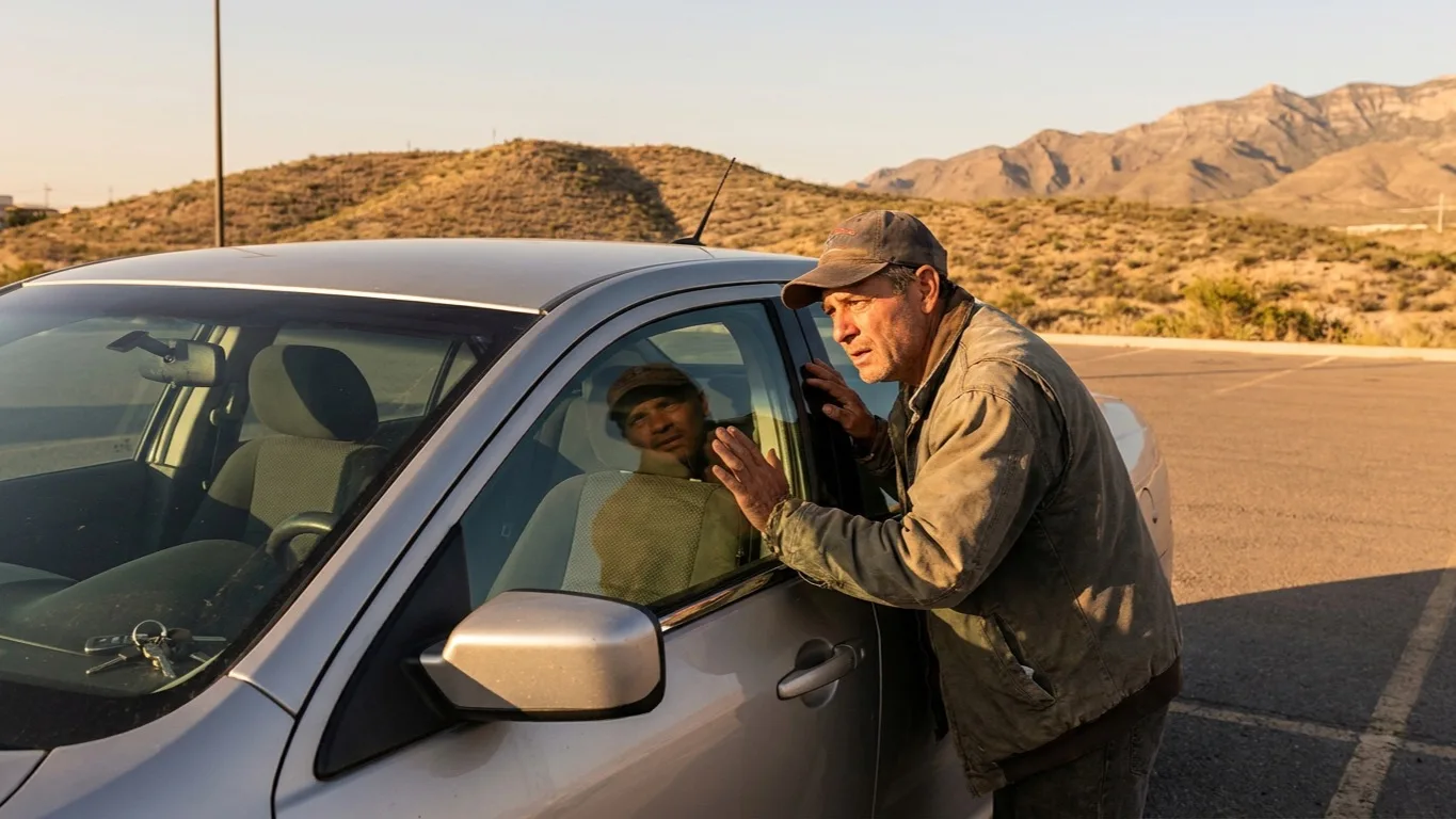 A worried driver peering through the window of a locked silver sedan in a desert El Paso parking lot at golden hour with the Franklin Mountains behind